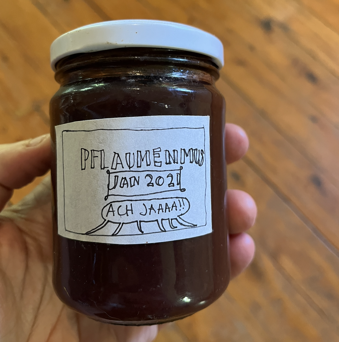 Close up of my hand holding a jar of jam, in front of a timber floor. The handwritten label reads "Pflaumenmus Jan 2021 Ach jaaaa!!"