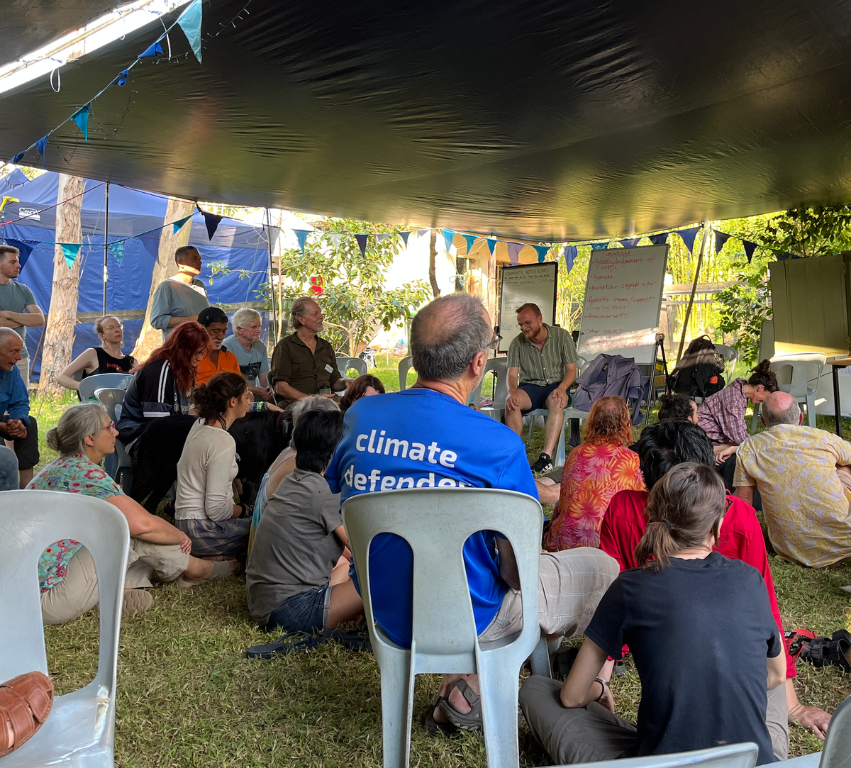 A group of people in a messy circle engaging in the "soft skill" of collaboration, under a tarpaulin on a grassy floor, with whiteboards