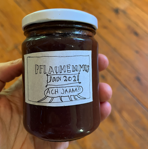 Close up of my hand holding a jar of jam, in front of a timber floor. The handwritten label reads "Pflaumenmus Jan 2021 Ach jaaaa!!"