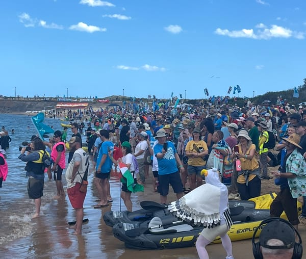 A performance activist in a seagull suit dances in front of the cops (out of shot), facing a HUGE crowd on people in colourful clothes on the beach, looking out at the water under a blue sky